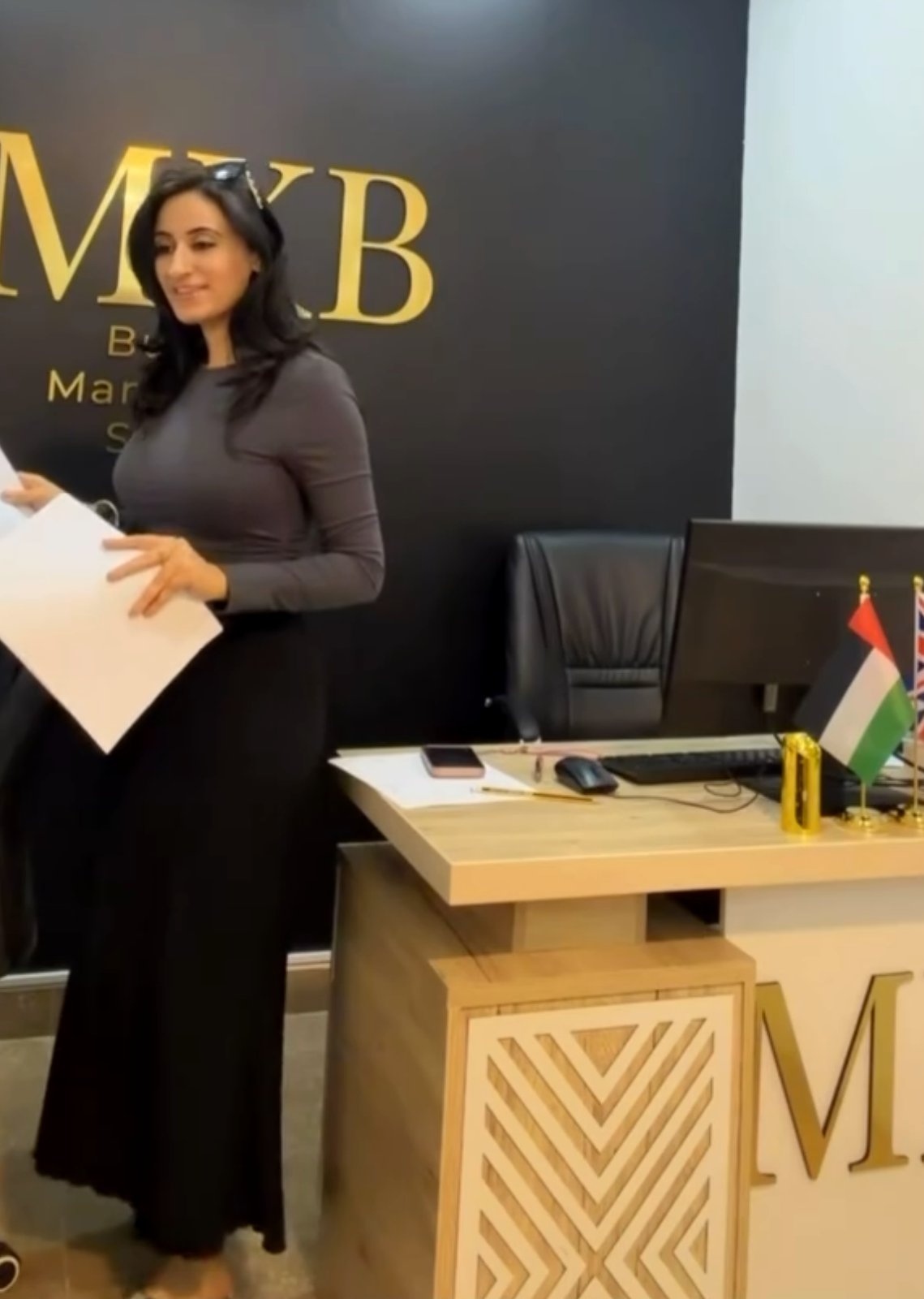 مهك بت مبتسمة وتحمل أوراق أحد العملاء أمام جدار شعار MKB الذهبي